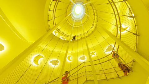 Blick nach oben in eine runde, gelb beleuchtete Kletterstruktur mit Netz und ausgeschnittenen Formen von Mond, Stern und Wolke, in der Kinder klettern
