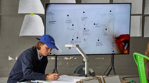 Person mit blauer Kappe sitzt an einem Tisch und zeichnet auf Papier vor einem Bildschirm mit handschriftlichen Notizen und Diagrammen