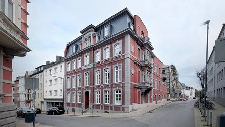 Ein historisches Eckgebäude mit roter Backsteinfassade, reich gegliederten Fensterrahmen und ausgebautem Dach steht an einer innerstädtischen Kreuzung, die sorgfältig sanierte Architektur fügt sich in die umliegende Blockstruktur ein, wodurch das Bild die behutsame Weiterentwicklung von Bestandsbauten zeigt und für Stadtreparatur, Identität und qualitätsvolle Nachverdichtung im urbanen Raum steht.