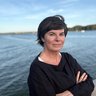 „Porträt einer Frau mit kurzen dunklen Haaren, die mit verschränkten Armen an einem sonnigen Tag vor einem weiten See steht.“