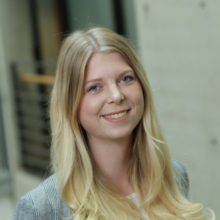 Porträt einer lächelnden Person mit langen blonden Haaren in Innenraumkulisse; unscharfer Hintergrund, graues kariertes Sakko sichtbar.