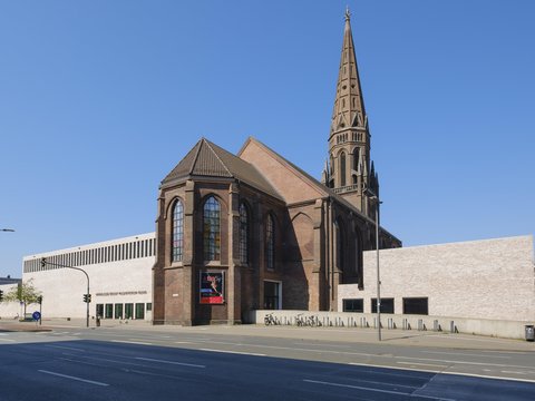 Außenansicht einer Backsteinkirche mit hohem, spitzem Turm; daran schließt ein moderner, heller Anbau mit schmalen Fensterbändern an. Breite Straße im Vordergrund, blauer Himmel.