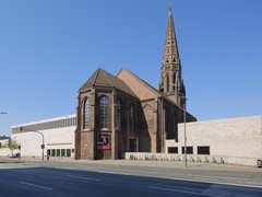 Außenansicht einer Backsteinkirche mit hohem, spitzem Turm; daran schließt ein moderner, heller Anbau mit schmalen Fensterbändern an. Breite Straße im Vordergrund, blauer Himmel.