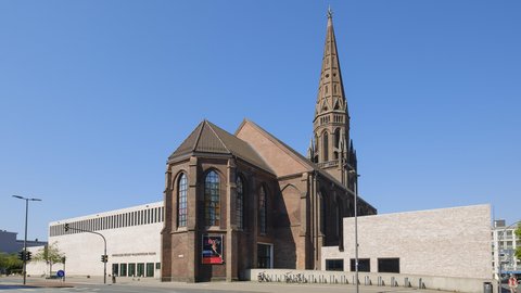 Außenansicht einer Backsteinkirche mit hohem, spitzem Turm; daran schließt ein moderner, heller Anbau mit schmalen Fensterbändern an. Breite Straße im Vordergrund, blauer Himmel.