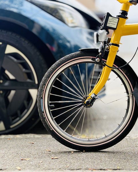 Vorderes Rad eines gelben Fahrrads vor einem geparkten Auto mit schwarzem Rad.