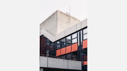 Gebäudedetail: helle Betonkante, schwarzes Fensterband und orangefarbene Brüstungsfelder; gläserner Erker links, heller Himmel.