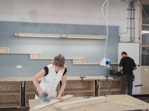 Ein dynamisches Foto aus einer geschäftigen Werkstatt. Im Vordergrund schleift eine Person mit hellen Haaren und Gehörschutz ein Holzbrett auf einer Hobelbank; ihre Bewegungen sind durch leichte Unschärfe eingefangen. Sie trägt ein weißes Oberteil. Im Hintergrund steht eine weitere Person in dunkler Kleidung und arbeitet an einer Werkbank, an der Schraubstöcke befestigt sind. Die Wand dahinter ist teilweise mit blauen OSB-Platten verkleidet und mit Holzregalboxen sowie einer langen Lichtleiste versehen. Werkzeuge und Kabel hängen von der Decke, und der Boden ist mit Holzspänen bedeckt. Die Atmosphäre ist produktiv und handwerklich.
