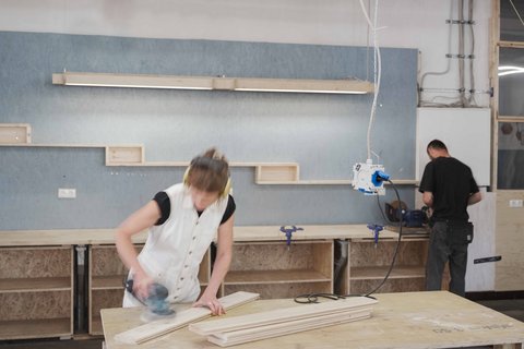 Ein dynamisches Foto aus einer geschäftigen Werkstatt. Im Vordergrund schleift eine Person mit hellen Haaren und Gehörschutz ein Holzbrett auf einer Hobelbank; ihre Bewegungen sind durch leichte Unschärfe eingefangen. Sie trägt ein weißes Oberteil. Im Hintergrund steht eine weitere Person in dunkler Kleidung und arbeitet an einer Werkbank, an der Schraubstöcke befestigt sind. Die Wand dahinter ist teilweise mit blauen OSB-Platten verkleidet und mit Holzregalboxen sowie einer langen Lichtleiste versehen. Werkzeuge und Kabel hängen von der Decke, und der Boden ist mit Holzspänen bedeckt. Die Atmosphäre ist produktiv und handwerklich.