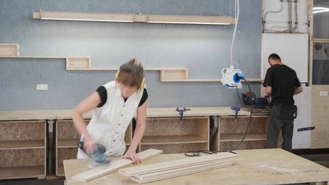 Ein dynamisches Foto aus einer geschäftigen Werkstatt. Im Vordergrund schleift eine Person mit hellen Haaren und Gehörschutz ein Holzbrett auf einer Hobelbank; ihre Bewegungen sind durch leichte Unschärfe eingefangen. Sie trägt ein weißes Oberteil. Im Hintergrund steht eine weitere Person in dunkler Kleidung und arbeitet an einer Werkbank, an der Schraubstöcke befestigt sind. Die Wand dahinter ist teilweise mit blauen OSB-Platten verkleidet und mit Holzregalboxen sowie einer langen Lichtleiste versehen. Werkzeuge und Kabel hängen von der Decke, und der Boden ist mit Holzspänen bedeckt. Die Atmosphäre ist produktiv und handwerklich.