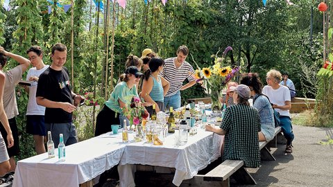Menschen sitzen und stehen an langen, weiß gedeckten Tischen in einem üppigen Garten. Über ihnen hängen bunte Wimpelketten, auf den Tischen stehen Blumen und Getränke