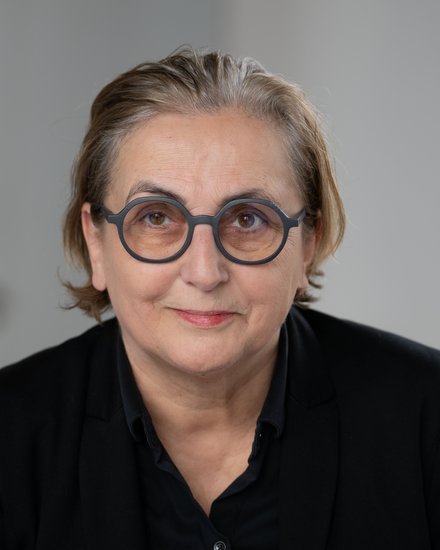 -Porträt einer Person mit schulterlangen, hellgrauen Haaren und runder Brille mit dunklem Rahmen. Sie trägt ein schwarzes Hemd und blickt direkt in die Kamera mit einem leichten Lächeln. Der Hintergrund ist weiß.