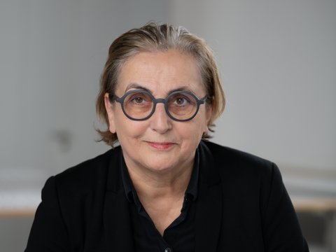 -Porträt einer Person mit schulterlangen, hellgrauen Haaren und runder Brille mit dunklem Rahmen. Sie trägt ein schwarzes Hemd und blickt direkt in die Kamera mit einem leichten Lächeln. Der Hintergrund ist weiß.