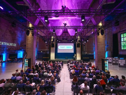 Blick von hinten in eine große, lilablau ausgeleuchtete Industriehalle: Viele sitzende Teilnehmende schauen auf eine Bühne mit Leinwand „THAT: 2025 – Digital planen. Mit Intelligenz gestalten.“ Ein Redner steht am Pult, über der Bühne hängen Lichttraversen; an den Seiten steht „ZENTRALHEIZE“.