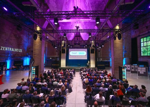 Blick von hinten in eine große, lilablau ausgeleuchtete Industriehalle: Viele sitzende Teilnehmende schauen auf eine Bühne mit Leinwand „THAT: 2025 – Digital planen. Mit Intelligenz gestalten.“ Ein Redner steht am Pult, über der Bühne hängen Lichttraversen; an den Seiten steht „ZENTRALHEIZE“.