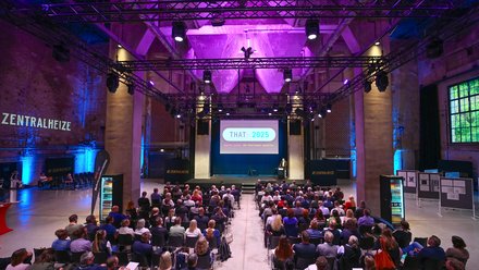 Blick von hinten in eine große, lilablau ausgeleuchtete Industriehalle: Viele sitzende Teilnehmende schauen auf eine Bühne mit Leinwand „THAT: 2025 – Digital planen. Mit Intelligenz gestalten.“ Ein Redner steht am Pult, über der Bühne hängen Lichttraversen; an den Seiten steht „ZENTRALHEIZE“.