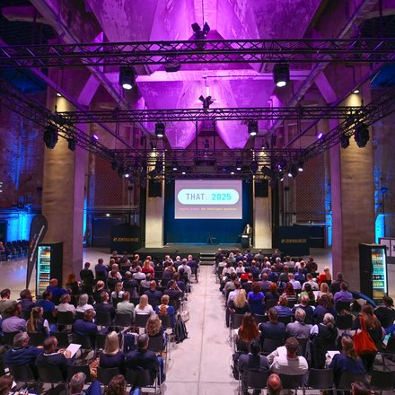 Blick von hinten in eine große, lilablau ausgeleuchtete Industriehalle: Viele sitzende Teilnehmende schauen auf eine Bühne mit Leinwand „THAT: 2025 – Digital planen. Mit Intelligenz gestalten.“ Ein Redner steht am Pult, über der Bühne hängen Lichttraversen; an den Seiten steht „ZENTRALHEIZE“.