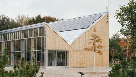 Holz‑ und Glasgebäude mit gezacktem Satteldach, Photovoltaik auf dem Dach und kleinem Windrad am Giebel; umgeben von Wiese und Bäumen.