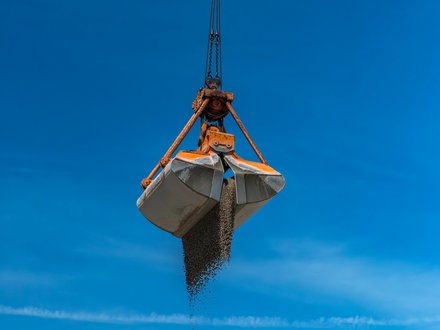 Foto Greiferkran Ein großer Greifer hängt am Kran und lässt Kies auf einen hohen Schüttkegel fallen; klarer blauer Himmel, Anlagengebäude im Hintergrund.