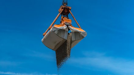 Foto Greiferkran Ein großer Greifer hängt am Kran und lässt Kies auf einen hohen Schüttkegel fallen; klarer blauer Himmel, Anlagengebäude im Hintergrund.