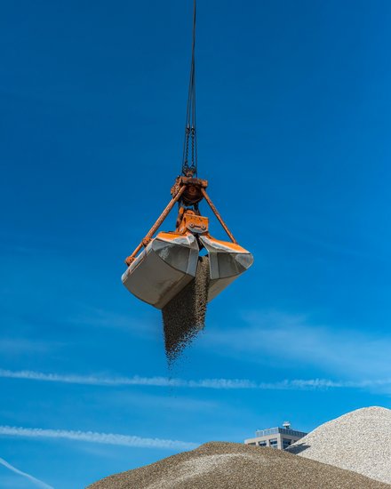 Foto Greiferkran Ein großer Greifer hängt am Kran und lässt Kies auf einen hohen Schüttkegel fallen; klarer blauer Himmel, Anlagengebäude im Hintergrund.