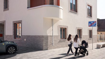 Eckgebäude mit weißer Fassade und abgerundeter Ecke, zwei Personen überqueren davor die Straße.