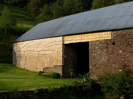 Ländliche Scheune mit Wellblechdach; Wände teils aus Bruchstein, teils mit hellen Holzschindeln verkleidet; davor eine grüne Wiese, dahinter Bäume.