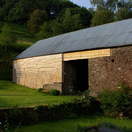 Ländliche Scheune mit Wellblechdach; Wände teils aus Bruchstein, teils mit hellen Holzschindeln verkleidet; davor eine grüne Wiese, dahinter Bäume.