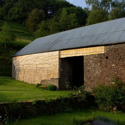 Ländliche Scheune mit Wellblechdach; Wände teils aus Bruchstein, teils mit hellen Holzschindeln verkleidet; davor eine grüne Wiese, dahinter Bäume.