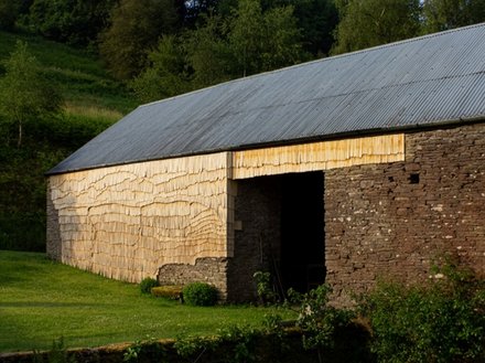 Ländliche Scheune mit Wellblechdach; Wände teils aus Bruchstein, teils mit hellen Holzschindeln verkleidet; davor eine grüne Wiese, dahinter Bäume.