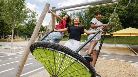 Drei Kinder stehen lachend auf einer großen Nestschaukel auf einem Spielplatz mit Bäumen und Sonnenschirm im Hintergrund