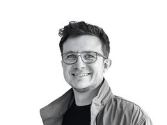 Schwarz-Weiß-Porträt eines lächelnden Philip Floegels mit Brille und Jacke vor weißem Hintergrund.