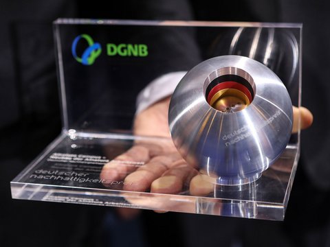 „Eine Person hält die Trophäe des Deutschen Nachhaltigkeitspreises der DGNB, bestehend aus einer gravierten Acrylplatte und einer Metallkugel, in den Händen.“