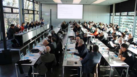 Großer Konferenzsaal mit vielen Teilnehmenden, die an langen Tischen sitzen und einer Präsentation auf der Leinwand folgen.