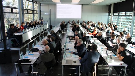 Großer Konferenzsaal mit vielen Teilnehmenden, die an langen Tischen sitzen und einer Präsentation auf der Leinwand folgen.