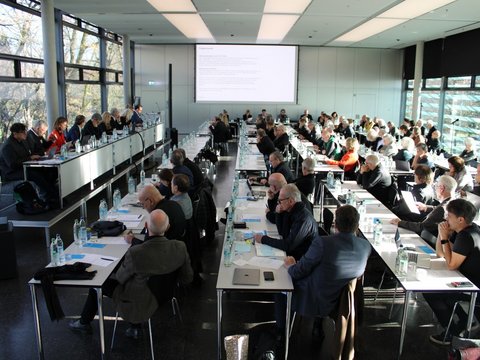 Großer Konferenzsaal mit vielen Teilnehmenden, die an langen Tischen sitzen und einer Präsentation auf der Leinwand folgen.