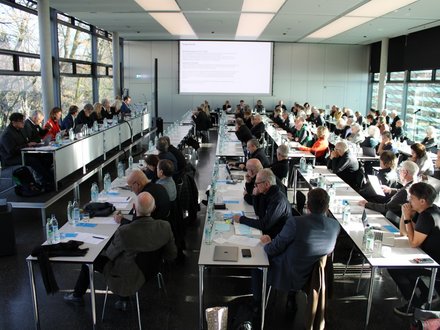 Großer Konferenzsaal mit vielen Teilnehmenden, die an langen Tischen sitzen und einer Präsentation auf der Leinwand folgen.