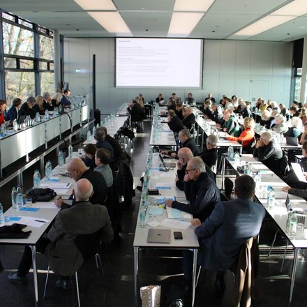 Großer Konferenzsaal mit vielen Teilnehmenden, die an langen Tischen sitzen und einer Präsentation auf der Leinwand folgen.
