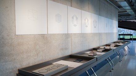 Ausstellung: weiße Tafeln mit schematischen Würfel‑Diagrammen an Betonwand; davor lange Tische mit Texten und Materialmustern.