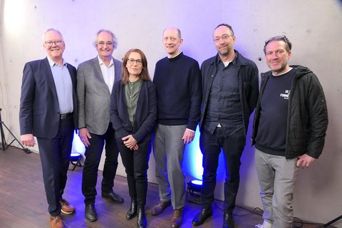 Sechs Personen stehen nebeneinander vor grauer Wand mit blauer Bodenbeleuchtung und blicken lächelnd in die Kamera; Gruppenfoto bei einer Veranstaltung.