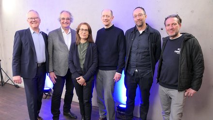 Sechs Personen stehen nebeneinander vor grauer Wand mit blauer Bodenbeleuchtung und blicken lächelnd in die Kamera; Gruppenfoto bei einer Veranstaltung.