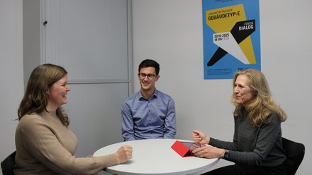 Drei Personen an rundem Tisch in einem Besprechungsraum: links eine Frau im beigen Pullover, in der Mitte ein junger Mann mit Brille, rechts eine ältere Frau mit Tablet‑Hülle; alle im Gespräch, Poster an der Wand im Hintergrund.