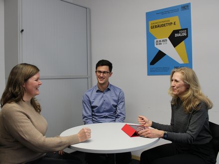 Drei Personen an rundem Tisch in einem Besprechungsraum: links eine Frau im beigen Pullover, in der Mitte ein junger Mann mit Brille, rechts eine ältere Frau mit Tablet‑Hülle; alle im Gespräch, Poster an der Wand im Hintergrund.