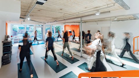 Gruppe von Personen beim Pilates-Training auf Matten in einem modernen Kursraum mit Spiegelfront, Betondecke und Bewegungsunschärfe.