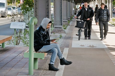 Mann mit Kapuzenjacke sitzt auf grüner Bank an Bahnsteig und liest Buch, im Hintergrund zwei Männer mit Fahrrad und Rucksack gehen