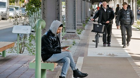 Mann mit Kapuzenjacke sitzt auf grüner Bank an Bahnsteig und liest Buch, im Hintergrund zwei Männer mit Fahrrad und Rucksack gehen