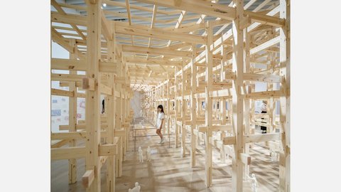 "Ausstellungsraum mit einer labyrinthartigen Struktur aus naturbelassenem Kiefernholz. Vertikale und horizontale Holzbalken bilden ein Rasterwerk, durch das Tageslicht fällt und geometrische Schatten wirft. An den Holzwänden sind Papierarbeiten, Skizzen und Ausdrucke befestigt. Eine Frau in weißem T-Shirt läuft durch das Labyrinth. Mehrere weiße Ausstellungsfiguren sind aufgestellt. Der Holzfußboden vervollständigt das helle, natürliche Ambiente."