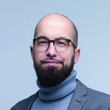 Porträtfoto: Person mit Glatze, Vollbart und Brille, in grauem Rollkragenpullover und grauem Sakko, schaut leicht lächelnd in die Kamera vor hellem, einfarbigem Hintergrund.