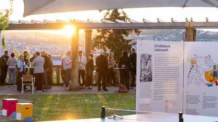 Abendliche Netzwerkveranstaltung im Freien: Menschen stehen unter einer Pergola mit Stadtblick im Sonnenuntergang; vorn Infotafel „(Um)Gebauten Lebenswelt“ und eine bunte Tischtennisplatte.