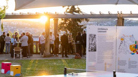 Abendliche Netzwerkveranstaltung im Freien: Menschen stehen unter einer Pergola mit Stadtblick im Sonnenuntergang; vorn Infotafel „(Um)Gebauten Lebenswelt“ und eine bunte Tischtennisplatte.