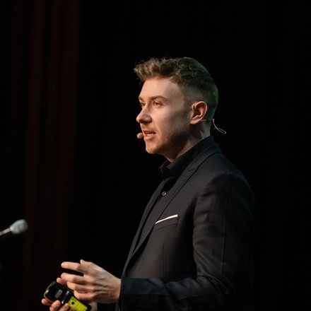 Vortrag eines Speakers auf dunkler Bühne Redner hält einen Vortrag mit Headset-Mikrofon vor schwarzem Hintergrund.